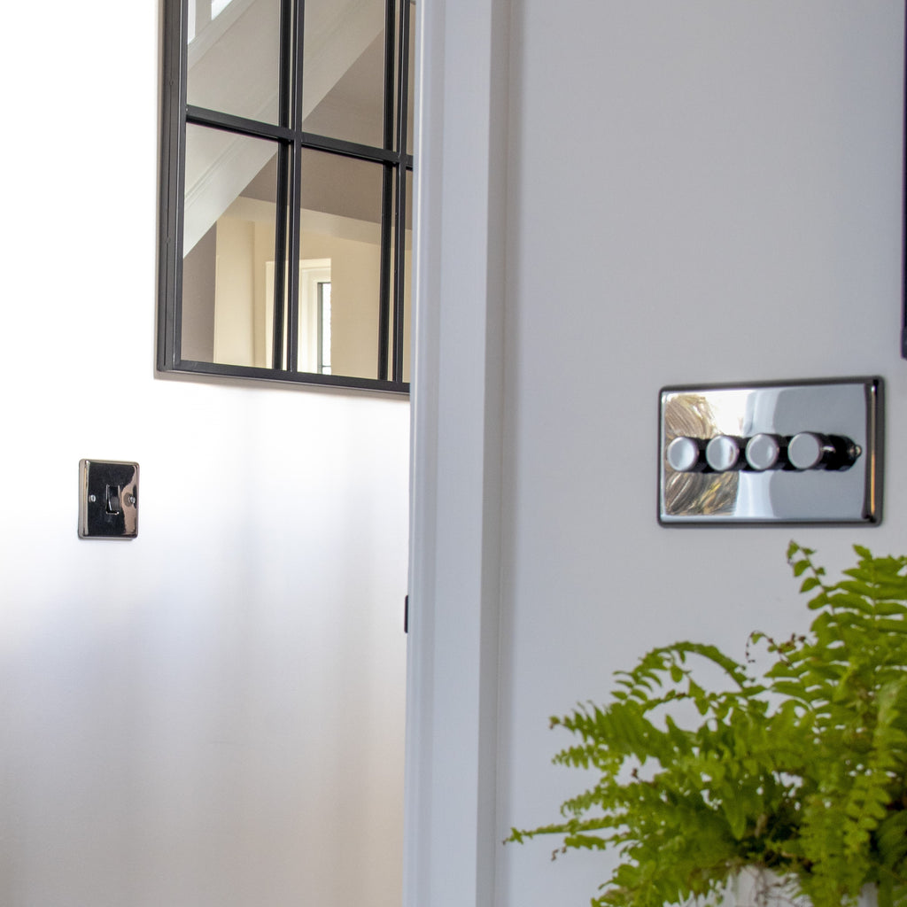 Chrome light switch with 4 switches on a white wall next to a door frame, with a hallway with a mirror in the background and a green fern in the corner.