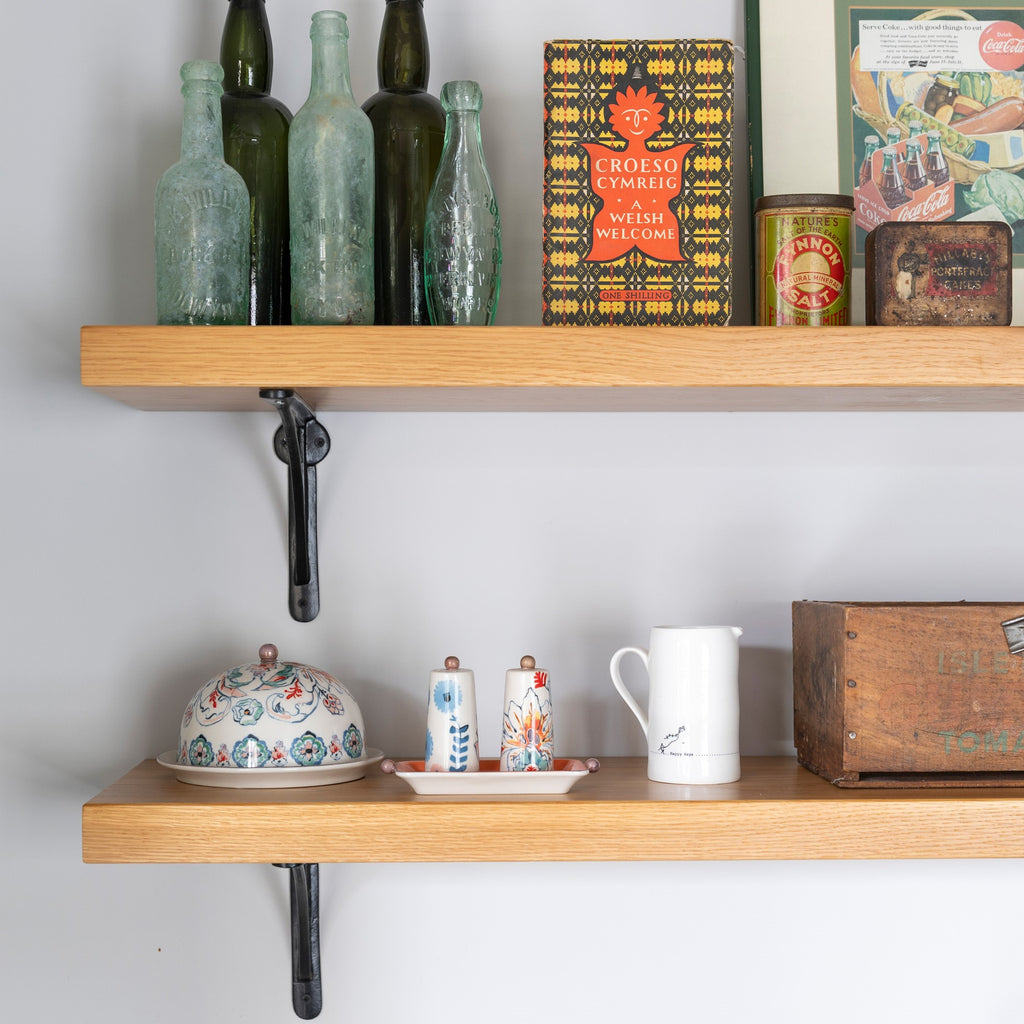 Black shelf brackets holding two wooden shelves filled with painted china, glass bottles and other knick-knacks