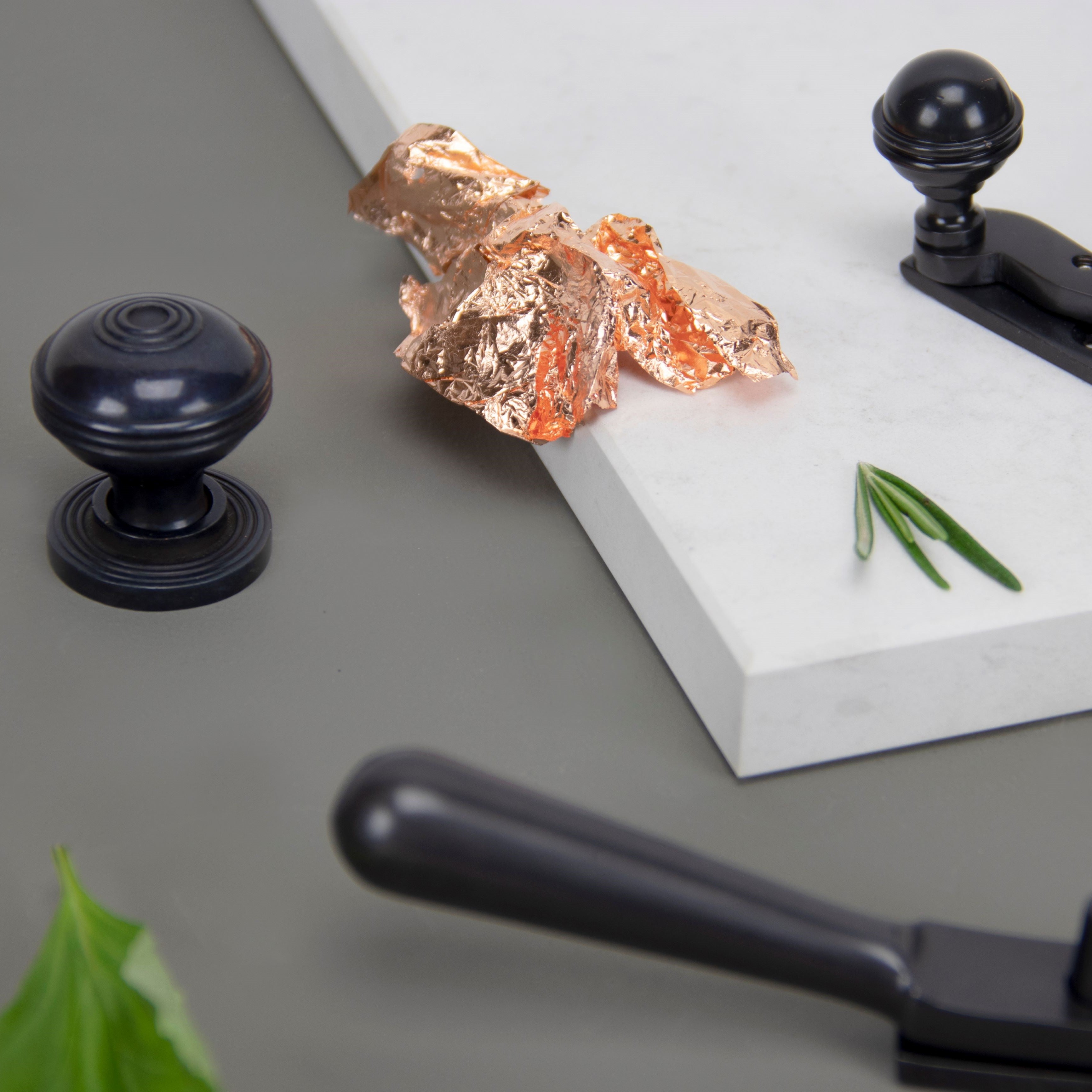 Ironmongery mood board with a grey background featuring our Aged Bronze window stay hardware and cabinet knob on a piece of white marble, with green leaves and gold foil.