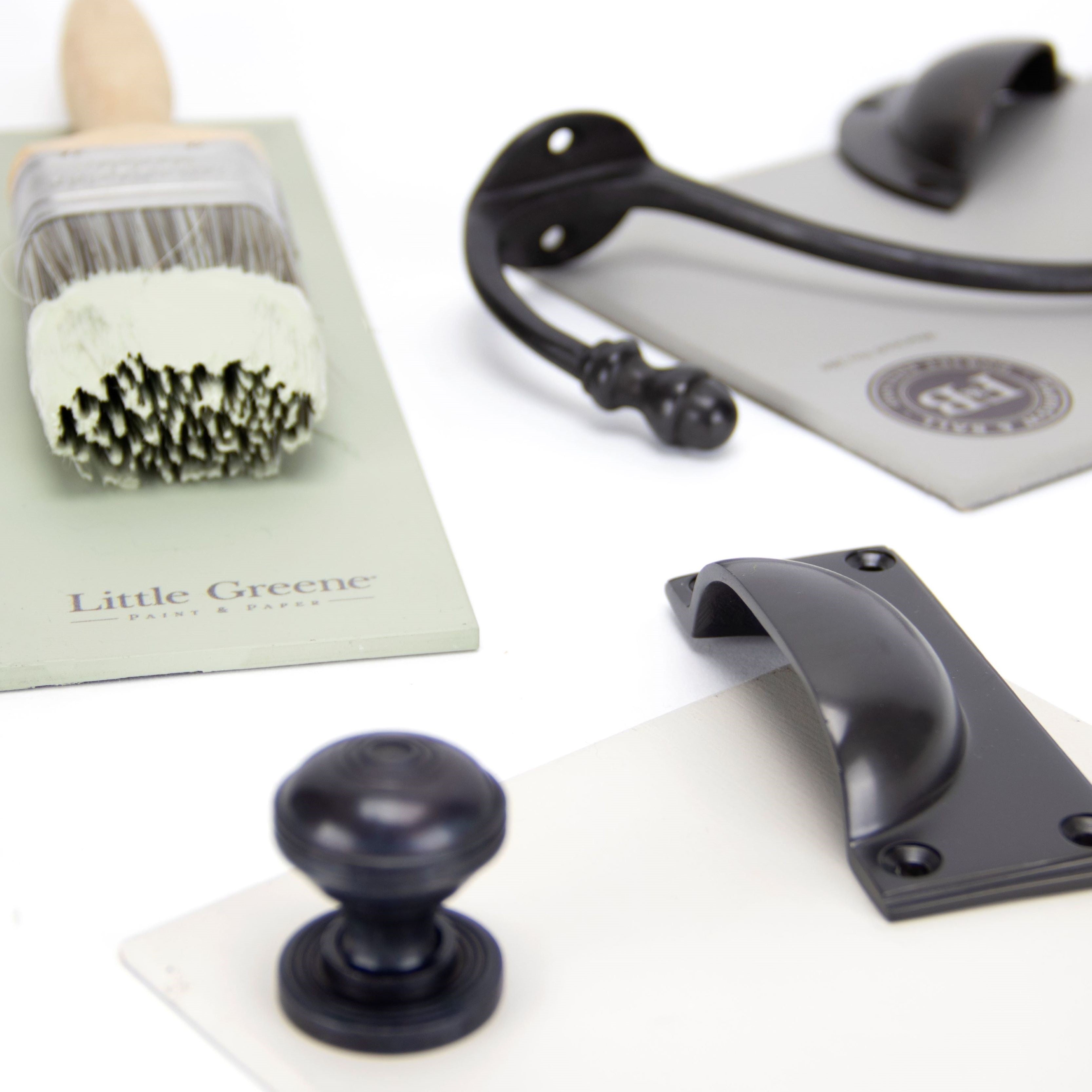 Ironmongery moodboard featuring our Aged Bronze coat hook, pull handle, drawer pull and cabinet knob, with Farrow & Ball paint samples and a wooden paintbrush with sage green paint.