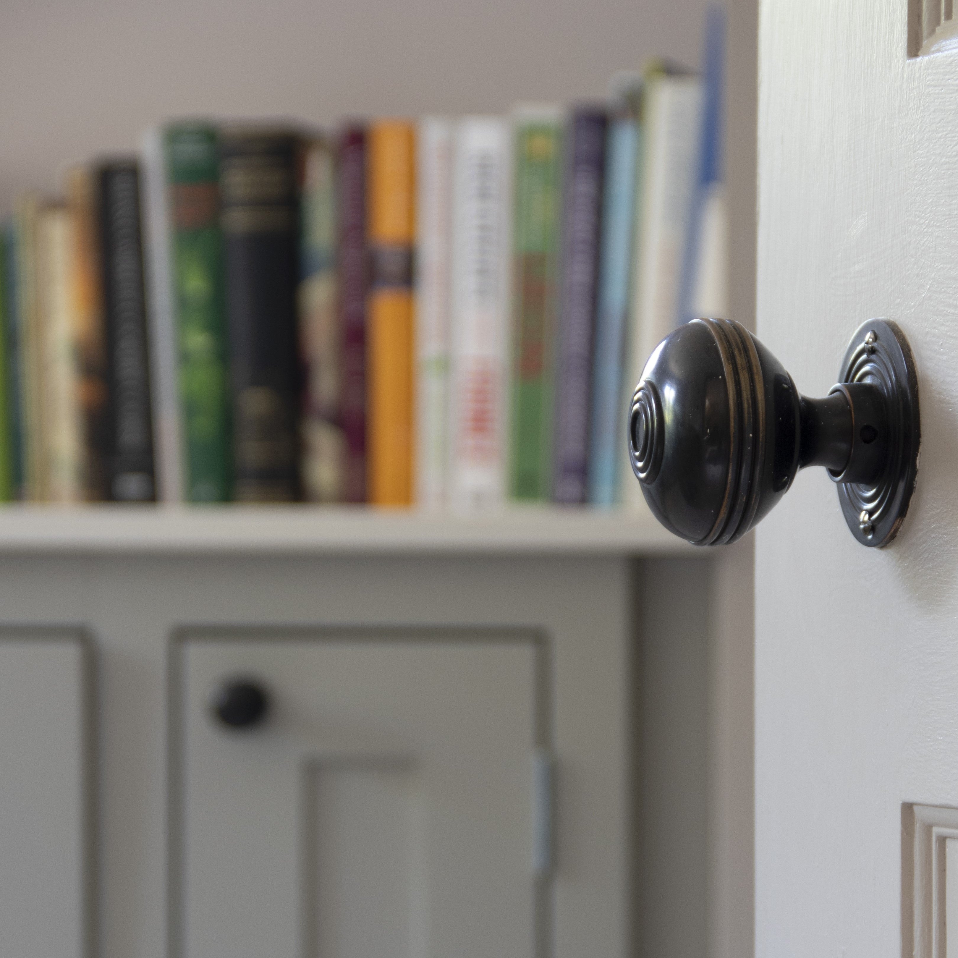 Bookshelf filled with books above a grey painted cupboard, with a panelled door with an Aged Bronze From The Anvil door knob in the foreground.
