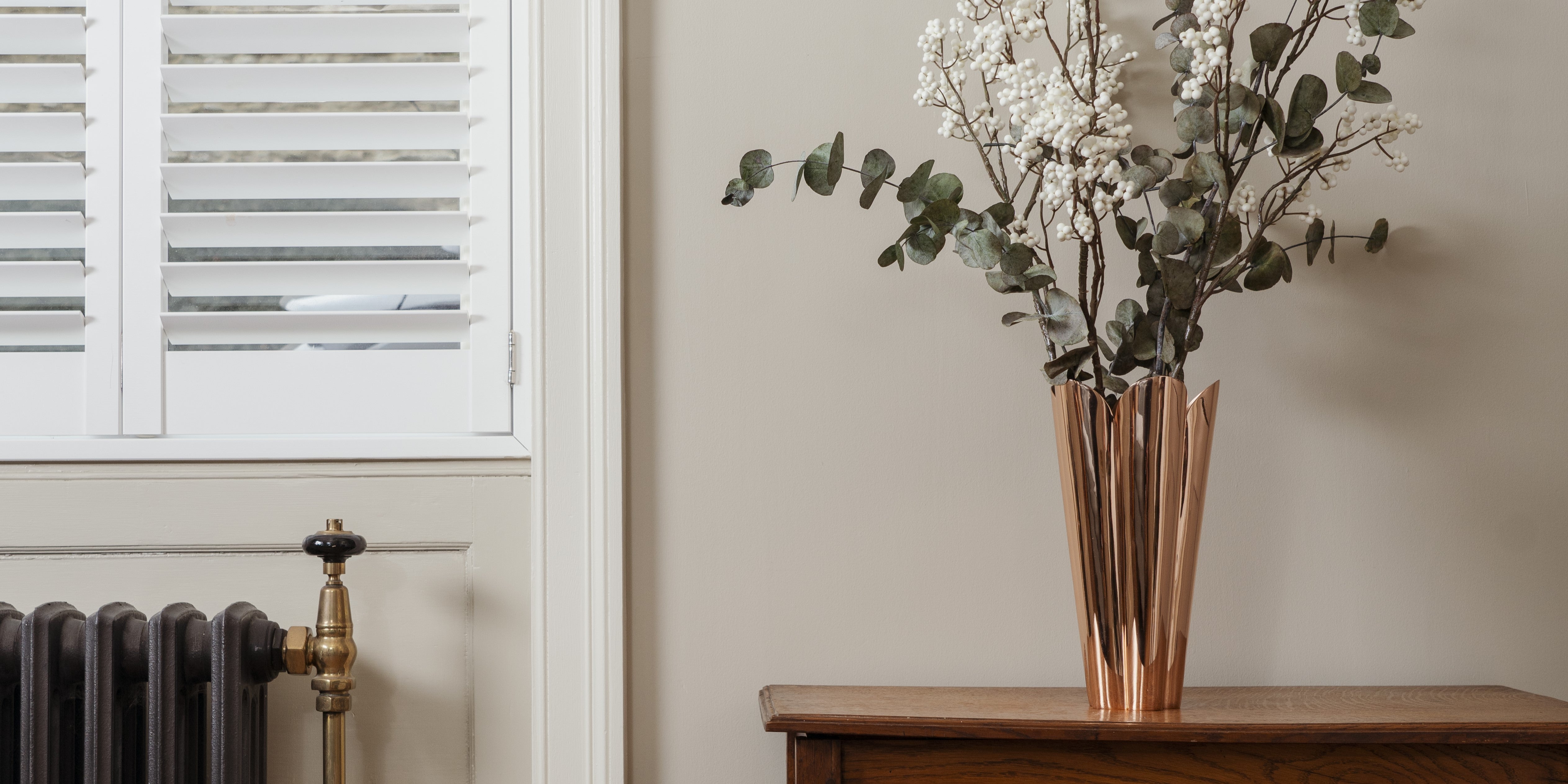 Smooth Copper Flora vase with a bouquet of flowers on a dark wood side table, next to a vintage radiator.