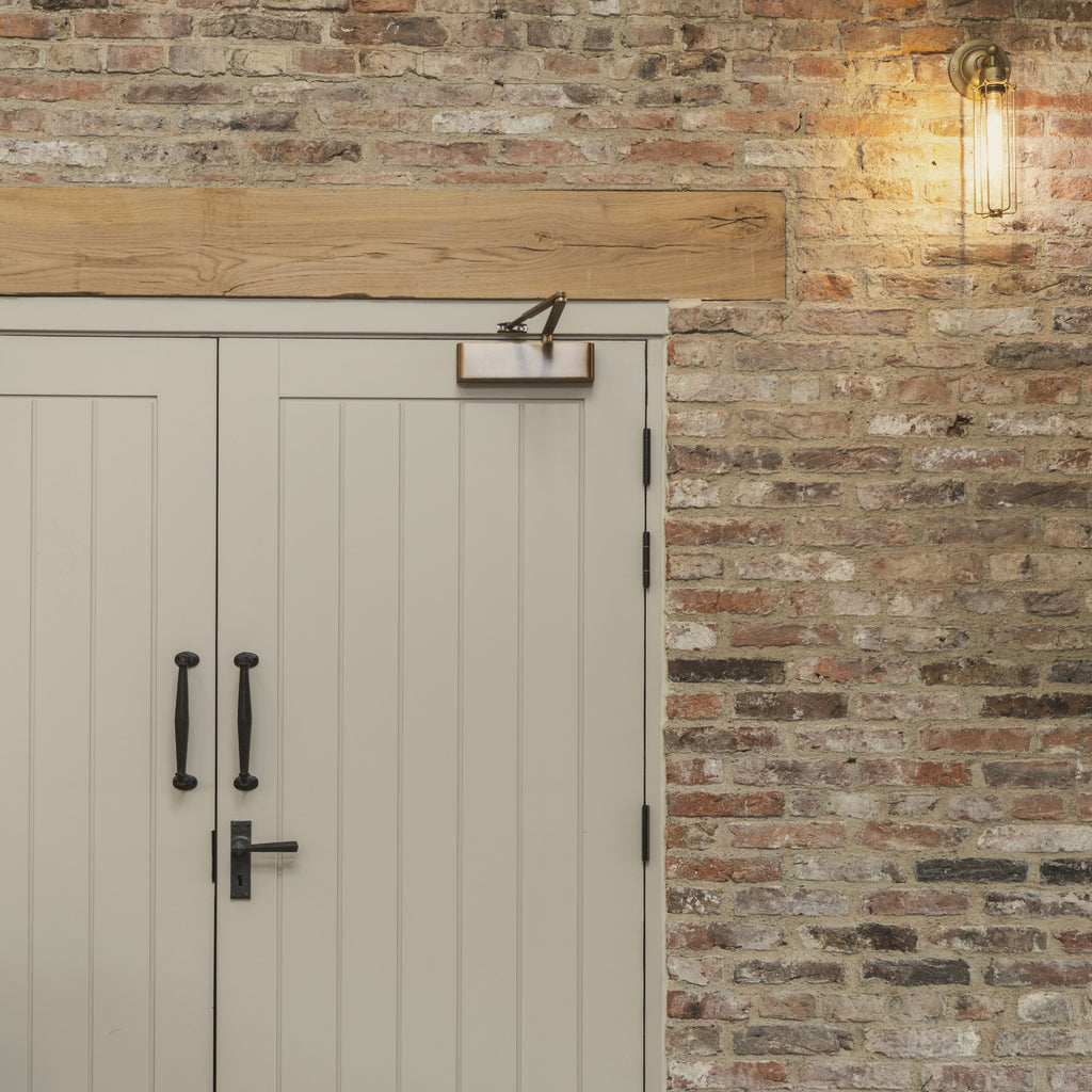 Painted double wooden doors with Beeswax pull handles and lever handle and Aged Brass door closer against a rustic brick wall.