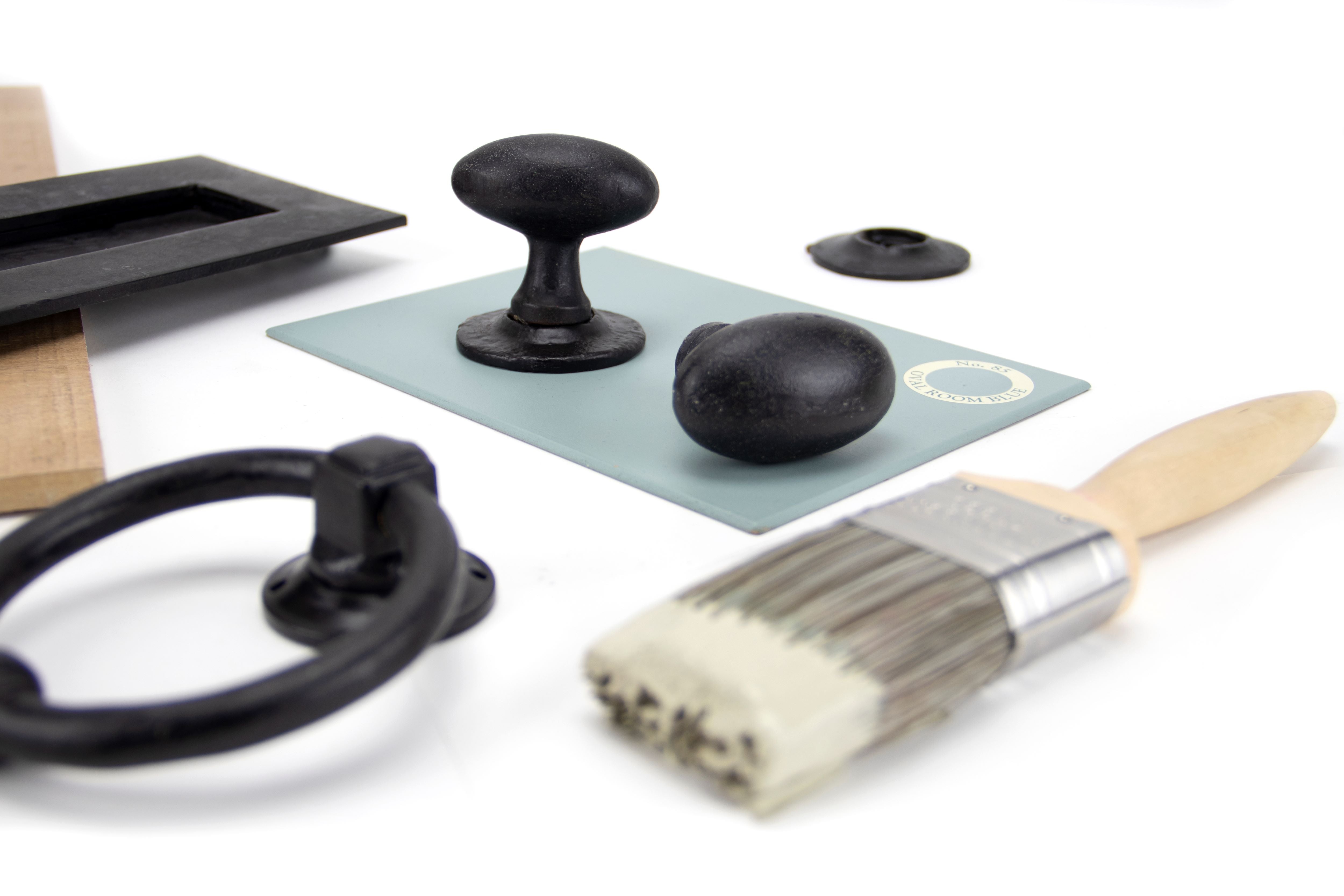 Ironmongery mood board with a white background featuring External Beeswax cabinet knobs, letterplate and door knocker on a blue paint swatch with a wooden paintbrush dipped in beige paint.