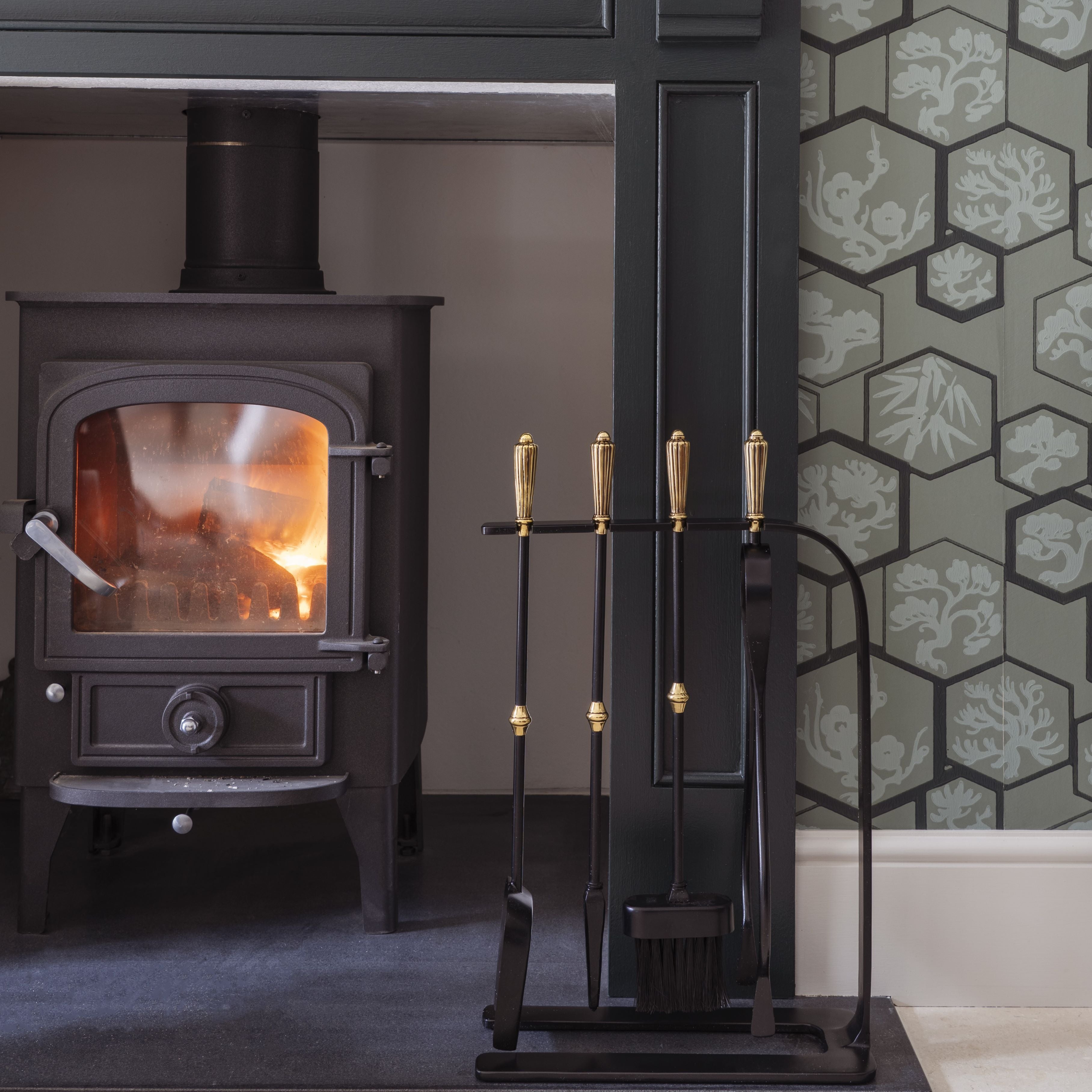 Black and Brass From The Anvil fireside tool companion set with tongs, brush, poker, and dustpan, next to a lit log burner.