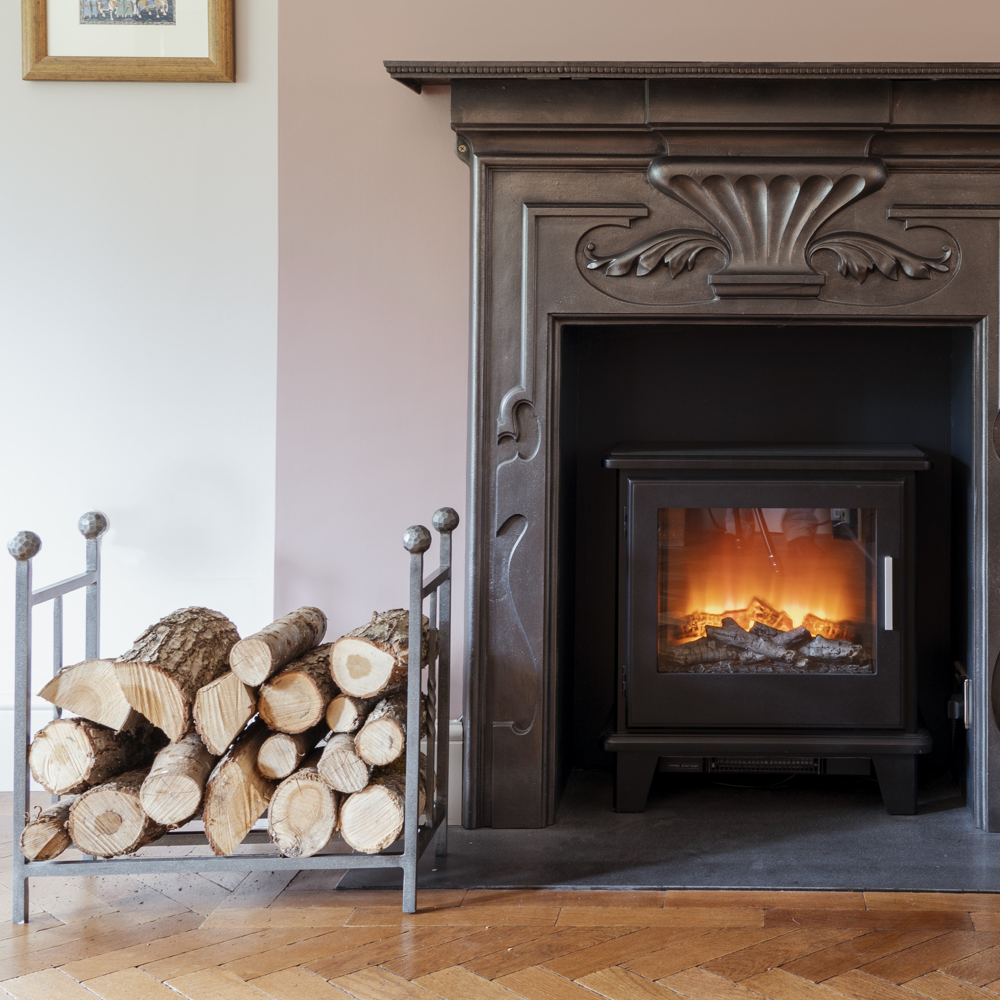 From The Anvil's Pewter Rectangular log holder filled with logs, next to a lit log burner within an antique fireplace.