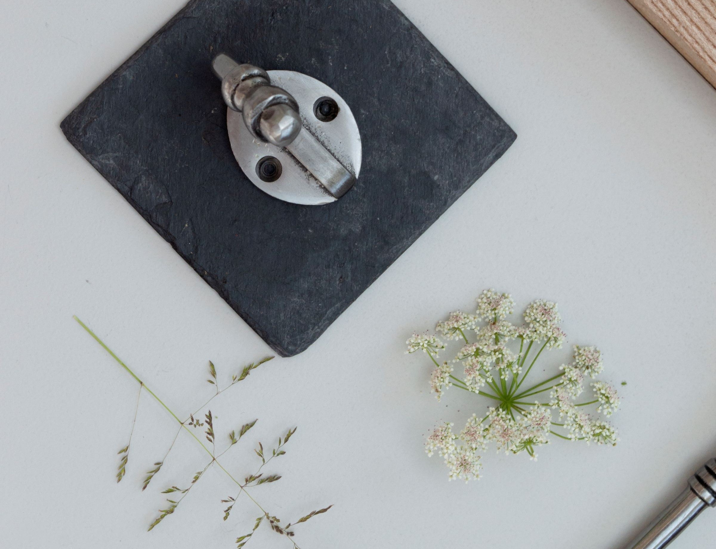 Pewter robe hook on small graphite slab on white background with dried flowers scattered around.
