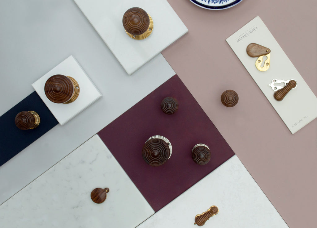 An ironmongery moodboard featuring rose wood cabinet handles, door knobs and escutcheons on various squares of marble, with burgundy, navy blue, and pale pink paint swatches in the background.