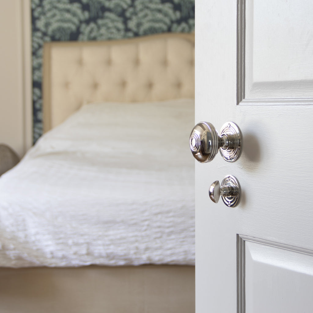 Polished Chrome door knob with matching thumbturn on a white panelled door with a cream bed in the background.