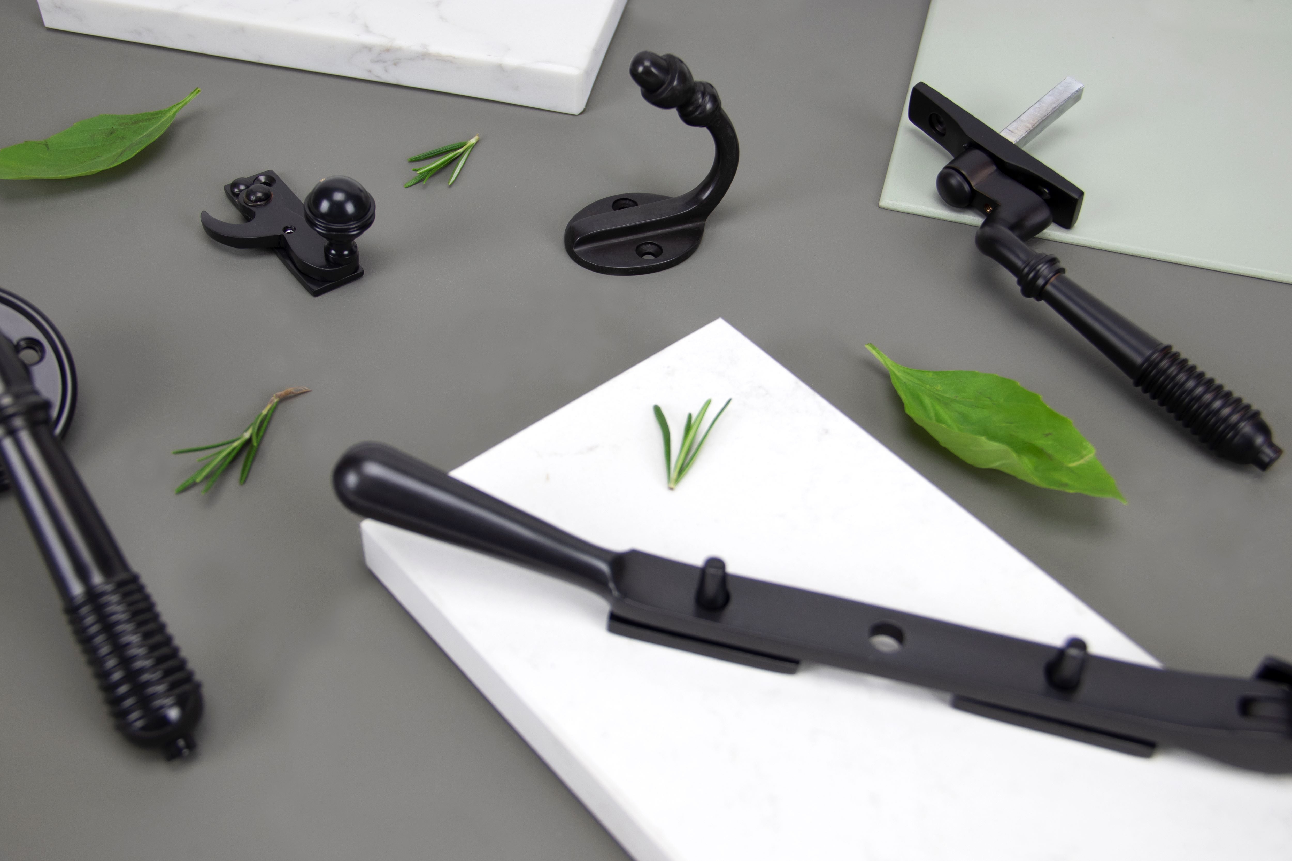 Ironmongery moodboard featuring Aged Bronze coat hook, window hardware and fastener, with a grey background, white marble slabs, and green leaves scattered around the products.