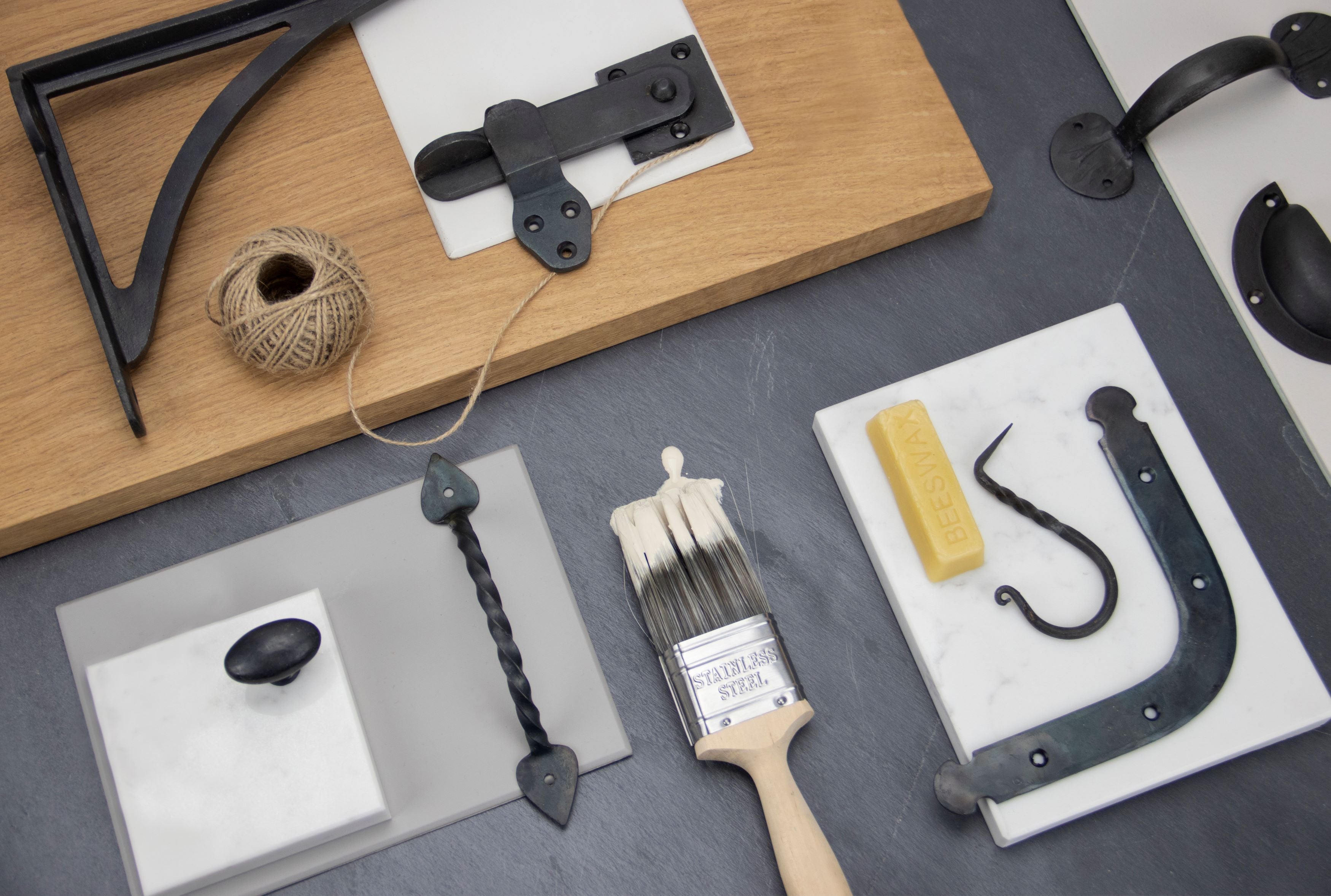 Ironmongery mood board with Beeswax shelf bracket, cabinet knob, pull handle and drawer pull, with a slab of wood, sheet of slate and some marble squares.
