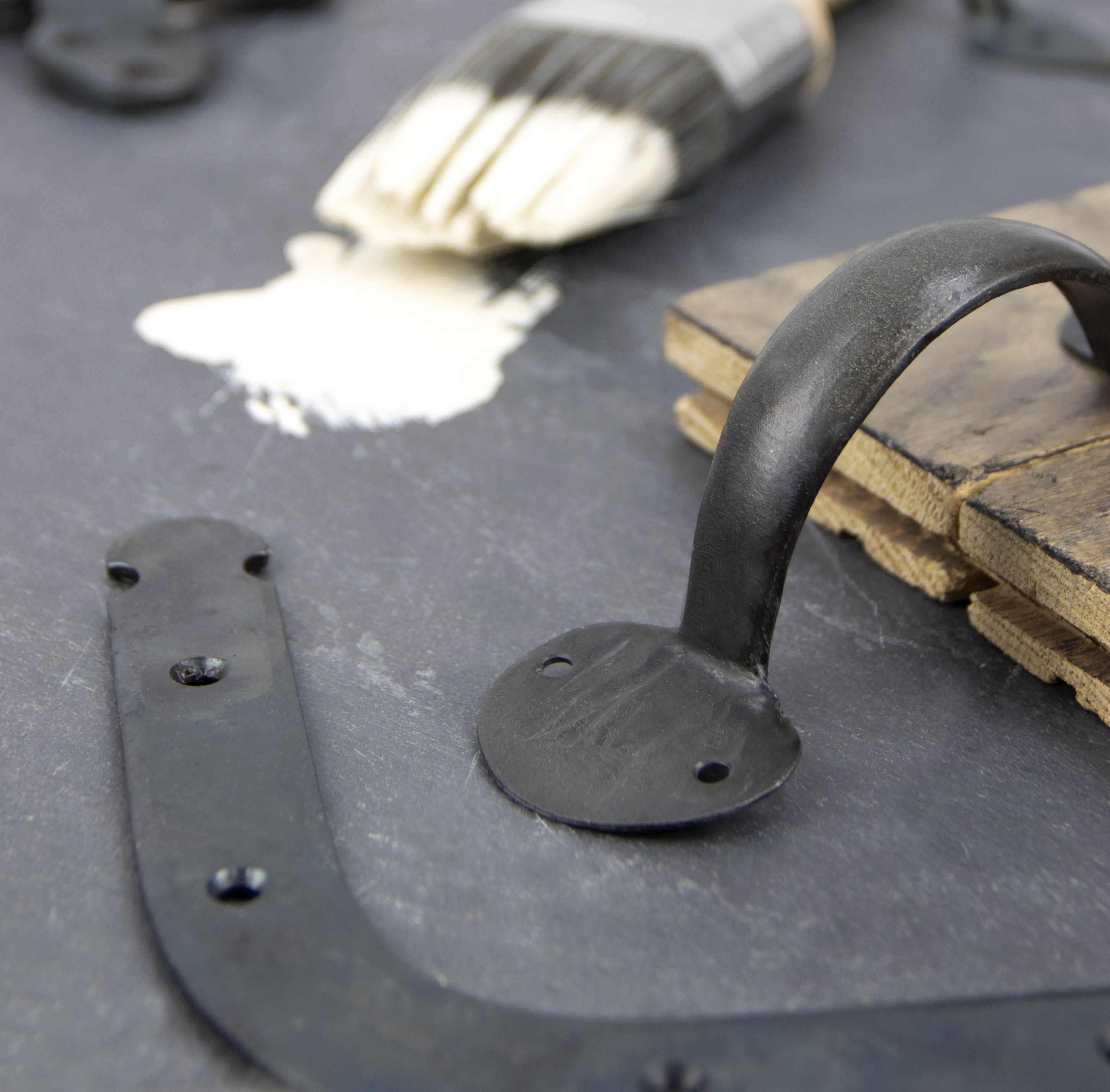 Ironmongery mood board on a sheet of slate, featuring our Beeswax shelf bracket and pull handles, and a wooden paint brush with cream Farrow & Ball paint on it.