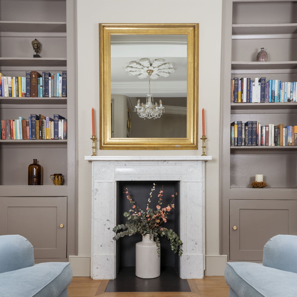 White marble fireplace with a gold framed mirror above and painted bookshelves on either side with From The Anvil Brass cabinet knobs,