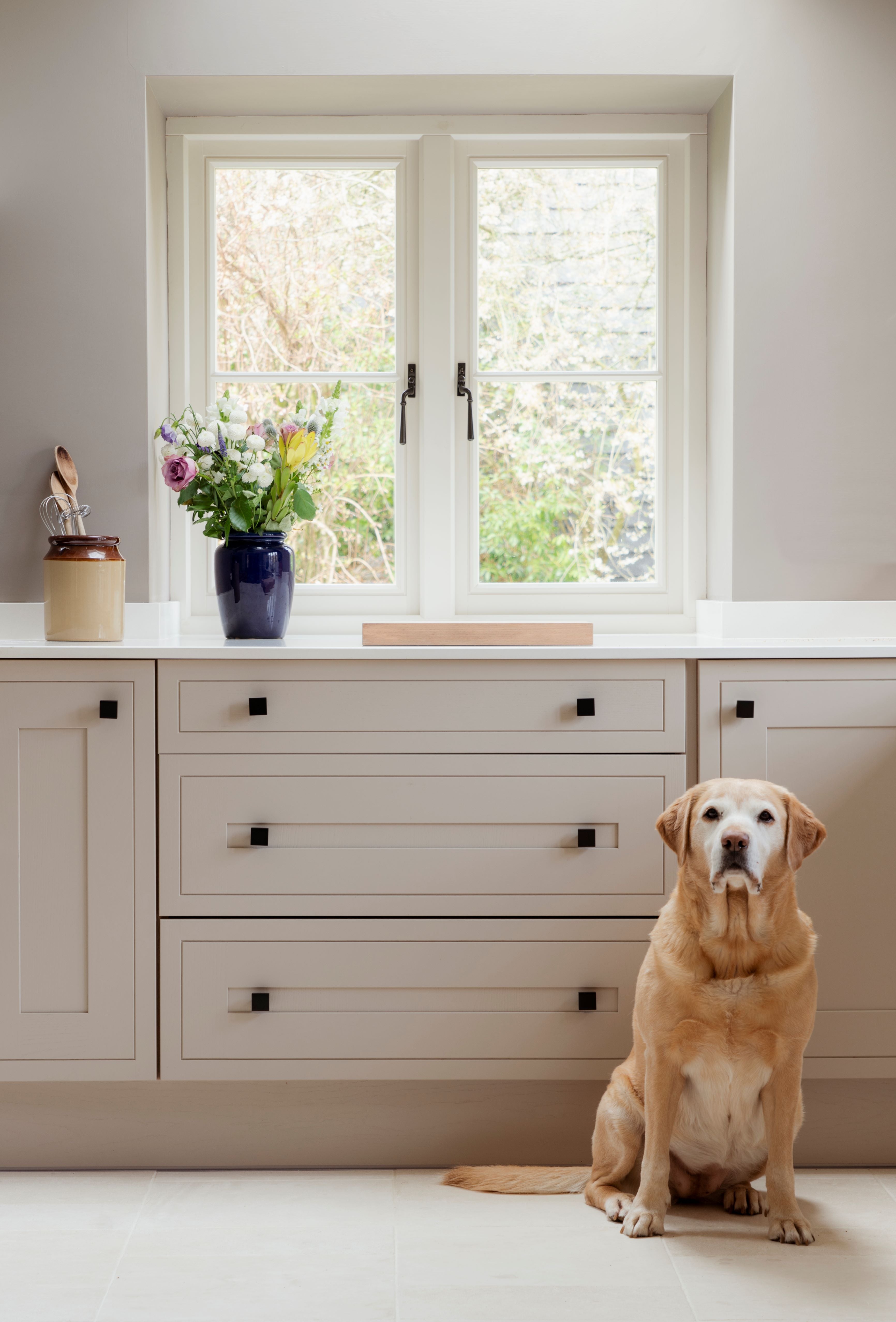 From The Anvil Aged Bronze window handles on a white uPVC window frame, and square cabinet knob handles on a cream kitchen below.