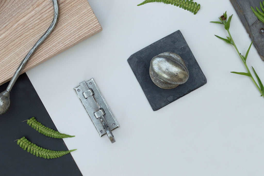 Pewter bolt, cabinet knob & hook on a white background with a piece of wood and green leaves.