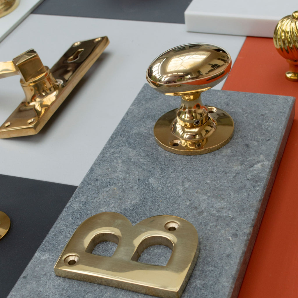 Polished Brass letter "B", cabinet knob and door handle on grey stone and orange and grey paint swatches.