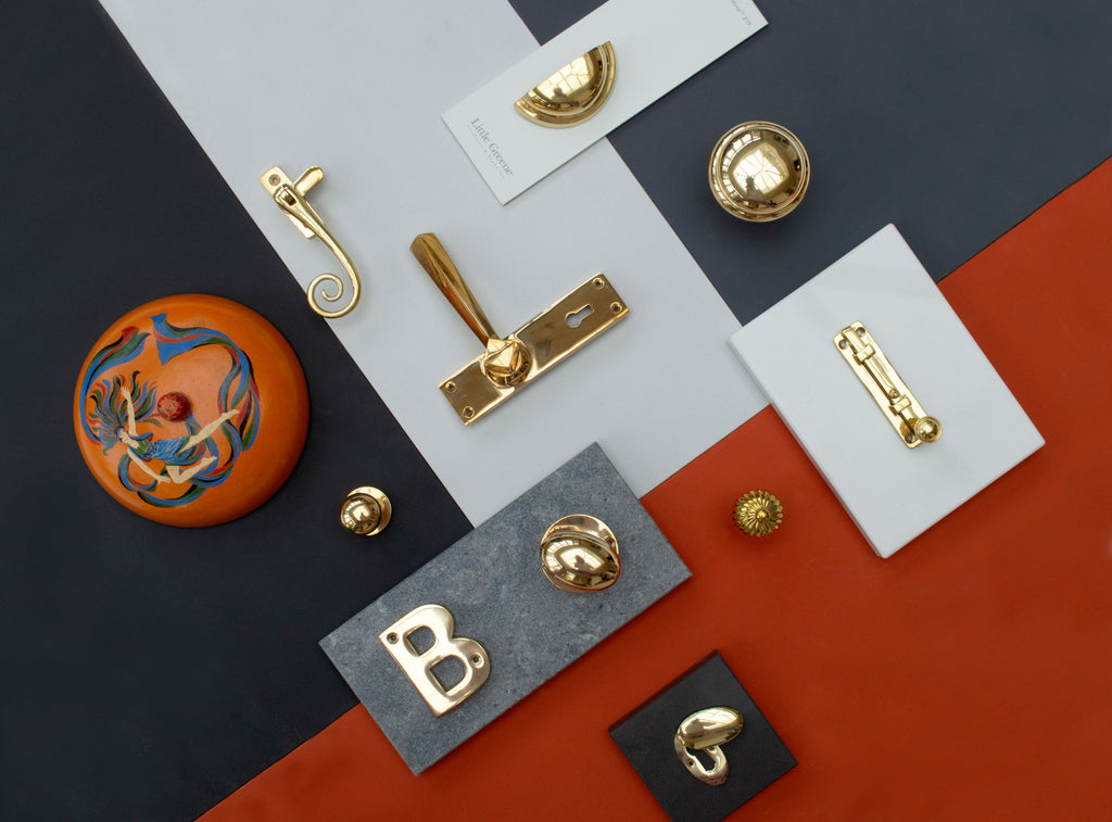 Ironmongery mood board with red, grey & navy background, featuring Polished Brass door handle, window hardware, door knob, cabinet knobs, letter "B", escutcheon and drawer pull, next to a decorative orange pot.