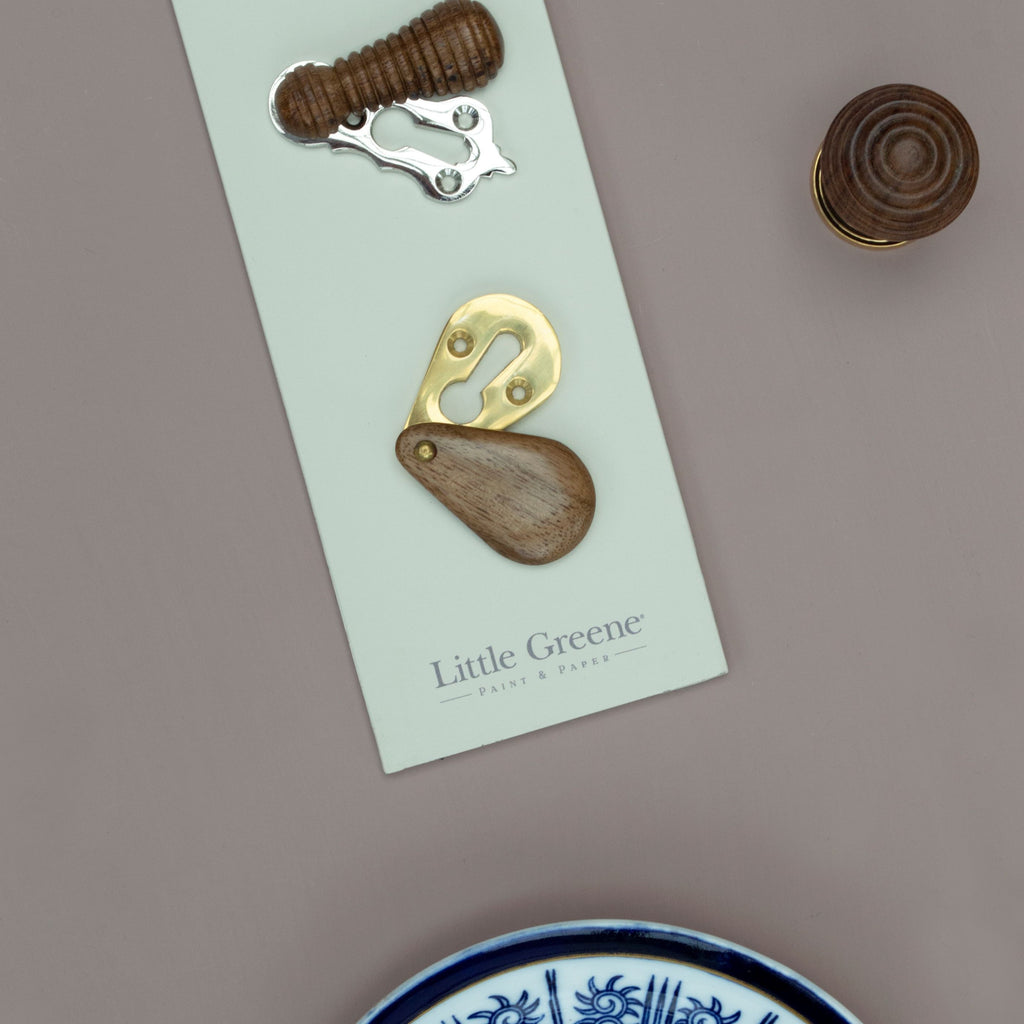 Wooden and Brass escutcheons on a pale green paint swatch with a wooden door knob in the top right corner, a blue and white lidded pot, and a pale pink background.